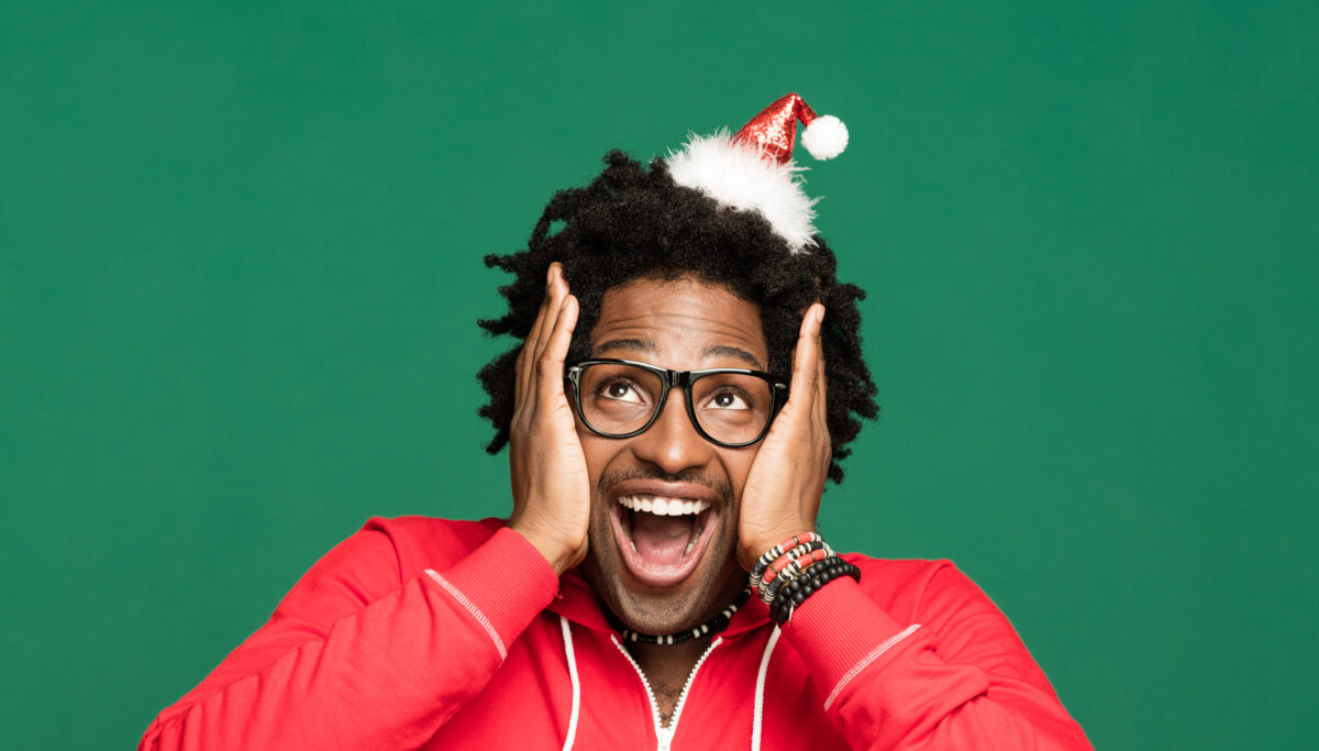 Funny Christmas portrait of young man wearing Santa Claus hat headband and red blouse, looking away and laughing. Studio shot against green background.