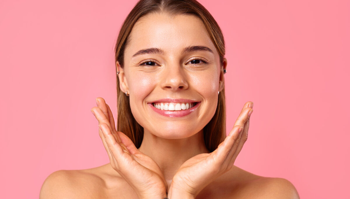 A cheerful young gently touching her face while posing against a pink background. The image conveys a theme of radiantly happy skin care