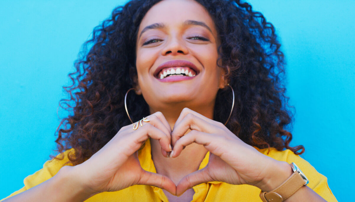 Heart, hands and portrait of woman with sign of love, care and empathy with a smile on blue, wall or studio background. Compassion, emoji and face of model with happiness in beauty or fashion