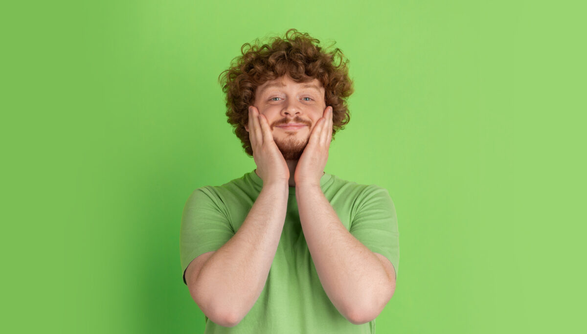 Shocked, astonished, wondered. Caucasian young man's monochrome portrait on green studio background. Beautiful male curly model in casual style. Concept of human emotions, facial expression.