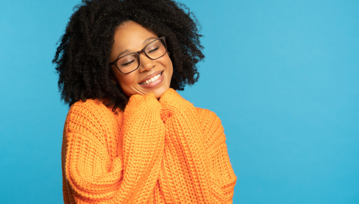 Stylish happy woman with curly hair wear orange knitted jumper, holds hands under chin , isolated on studio blue background. Winter warm clothes.