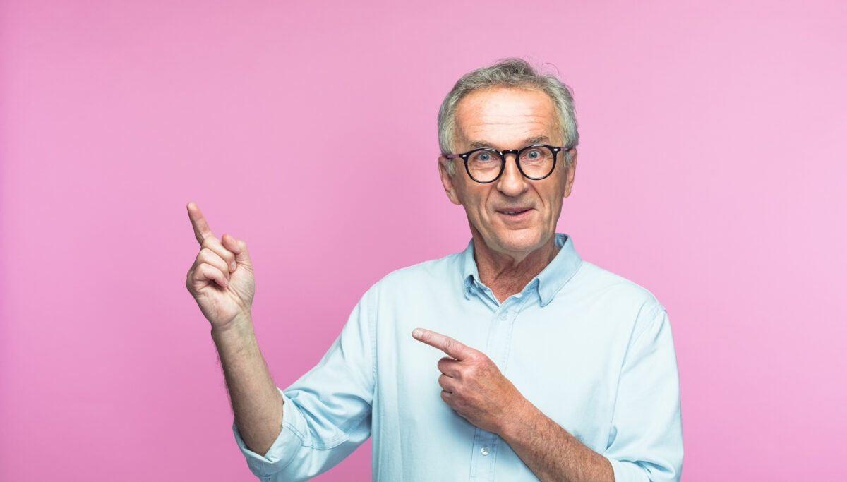 Portrait of smiling senior man pointing at copy space while standing against pink background
