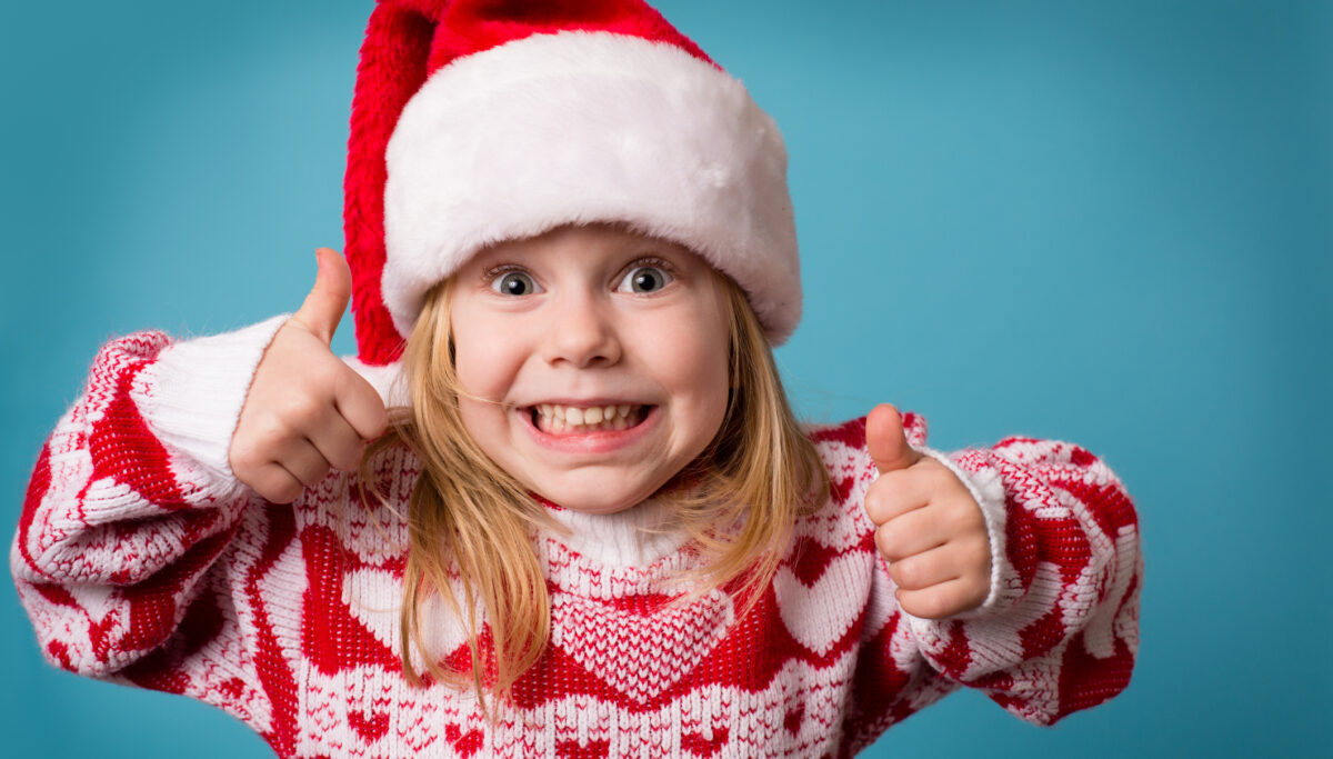 "Color image of a little girl wearing a Santa hat and an ugly Christmas sweater, smiling and making the thumbs up sign."