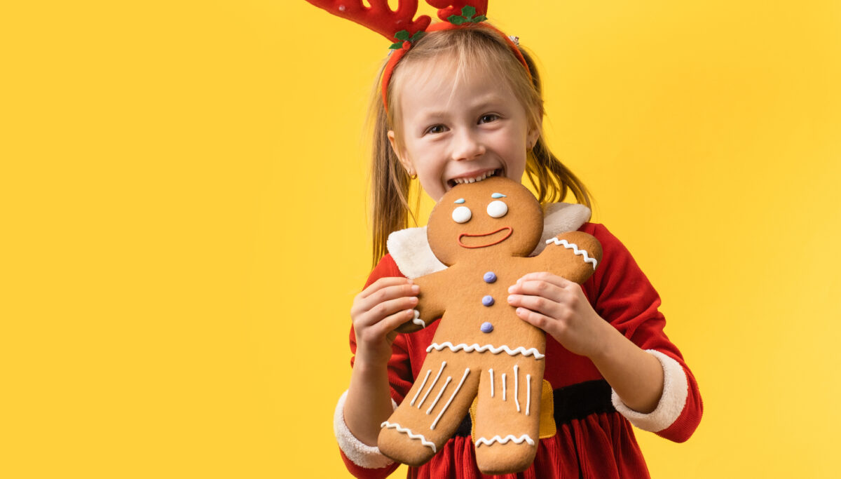 Cute little girl wearing Santa red costume and holding big baked gingerbread man cookie on the yellow background. Portrait of a happy smiling 5 year old girl on the Christmas background