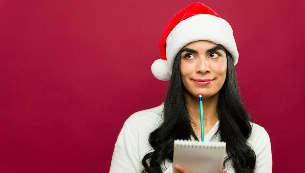 Thoughtful woman wearing a santa hat holding a notepad and making her christmas shopping list while smiling