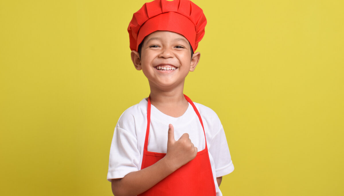 Cute little chef showing thumbs up isolated on yellow background. Asian boy wearing red apron and hat. Great little chef