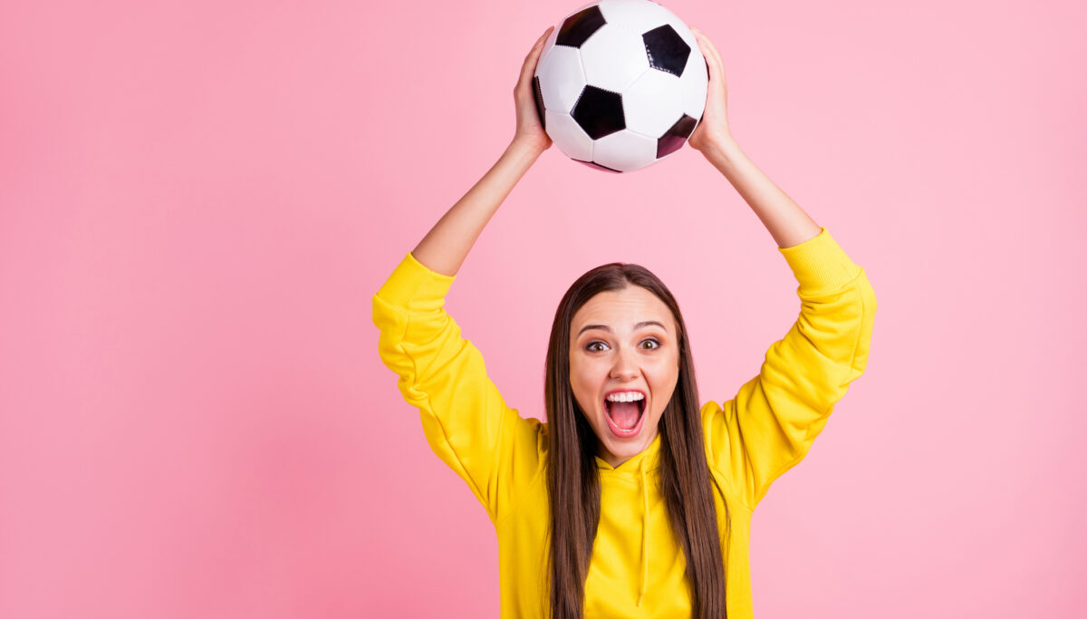 Photo of charming overjoyed girl holding soccer ball above her, head wearing yellow sweater while isolated over pink pastel color background