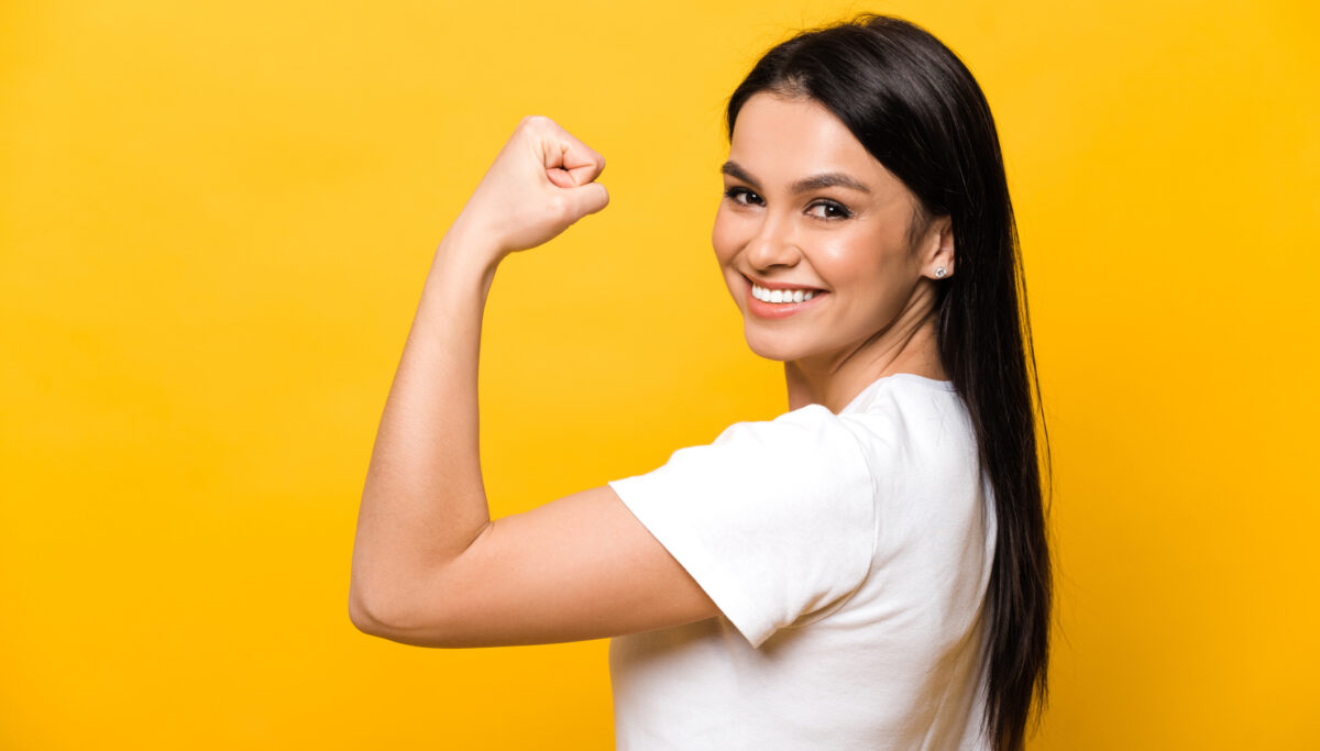 Happy and smiling woman standing showing her strength using her arm against an orange background.