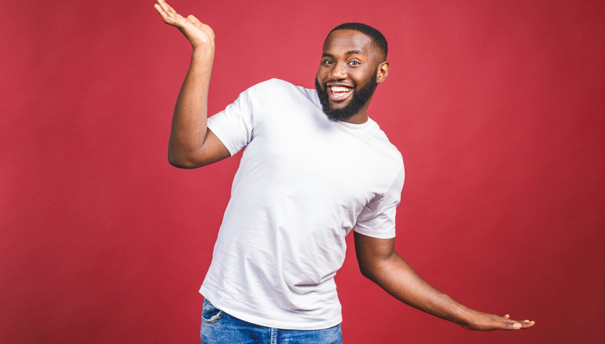 Funny guy in white t-shirt jumping and looking at camera. Studio portrait of emotional african male model posing on red background.
