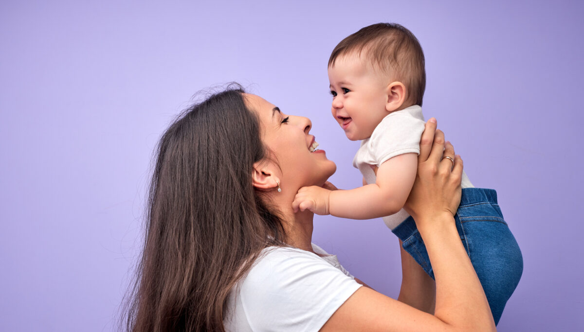 Beautiful young caucasian mother tossing up child baby. Baby laughing. Purple background. In studio. Copy space, side view. Portrait of caucasian family. Happy childhood concept
