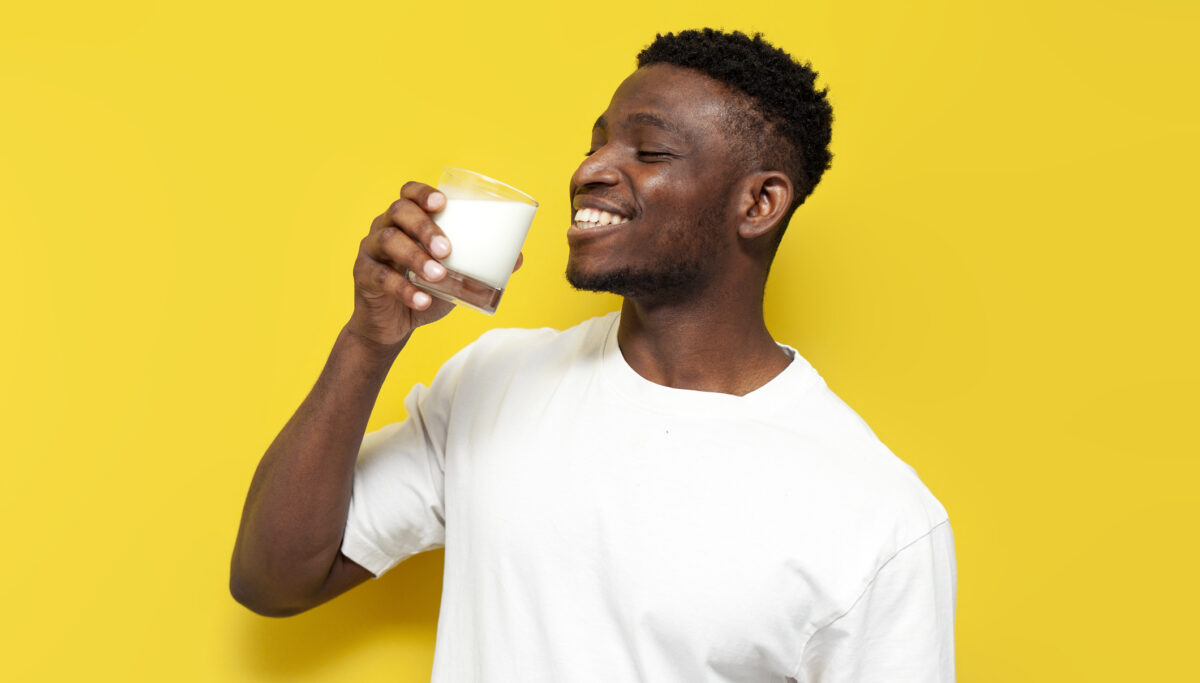 Man in white t-shirt drinks milk in glass on yellow isolated background, the guy holds glass of fresh kefir and smiles