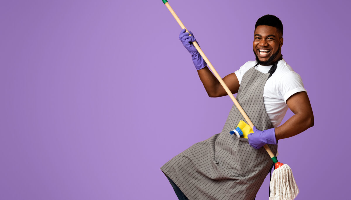 Portrait Of Funny Man In Apron With Mop In Hands Posing Over Pink Studio Background, Using Handle As Guitar, Fooling With Household Tool