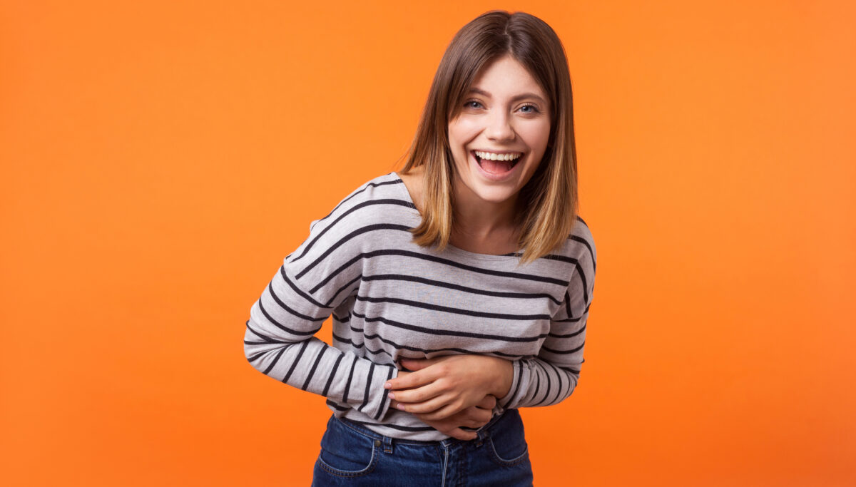 Portrait of overjoyed excited adorable young woman with brown hair in long sleeve striped shirt standing, holding her belly and laughing out loud. indoor studio shot isolated on orange background