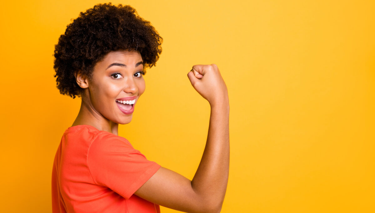 A happy woman, flexing her arm muscle, on a yellow coloured background.