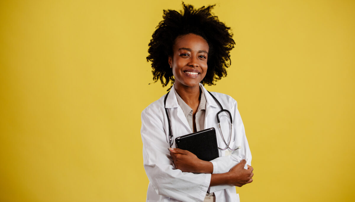 Young female doctor/scientist in white lab coat holding digital tablet and smiling while standing against yellow background.