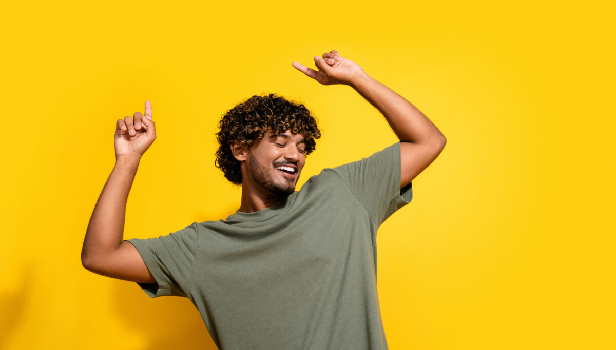 Photo of carefree good mood guy with wavy hairstyle dressed khaki t-shirt dancing raising fingers up isolated on yellow color background.