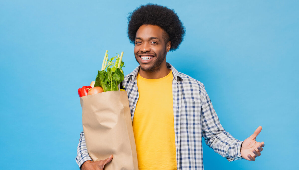 Friendly smiling Afro African American man holding paper shopping bag full of groceries in light blue isolated studio shot background