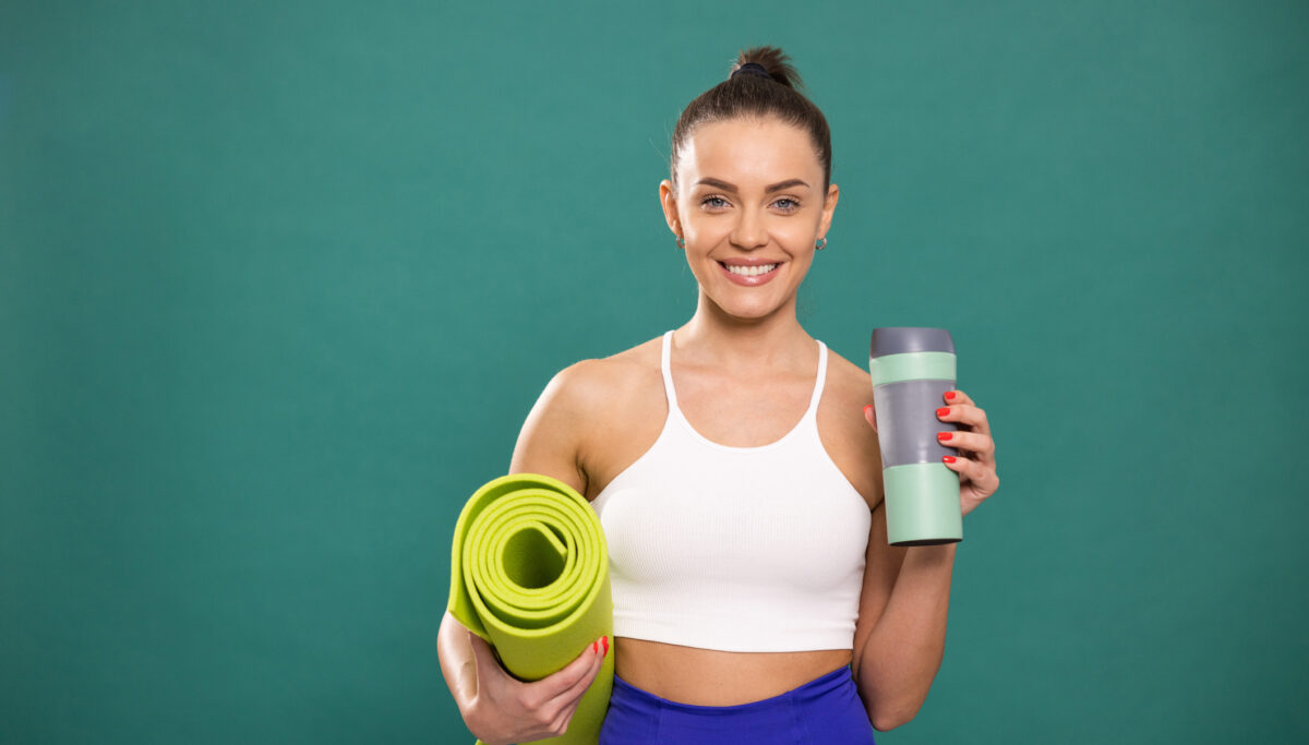 Young smiling woman standing in studio in front of green background while holding exercise mat and water bottle and looking at camera