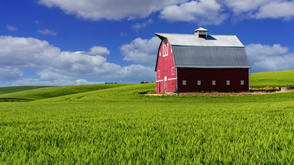 A photo of a barn, surrounded by green fields and blue skies