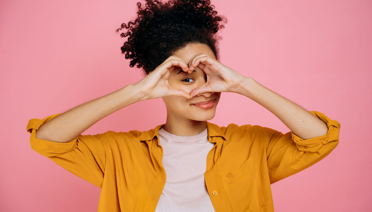 Lovely candid pleasant girl, makes heart gesture with hands near eye, demonstrates love sign, stands on isolated pink background, looking at camera though love gesture, smile friendly