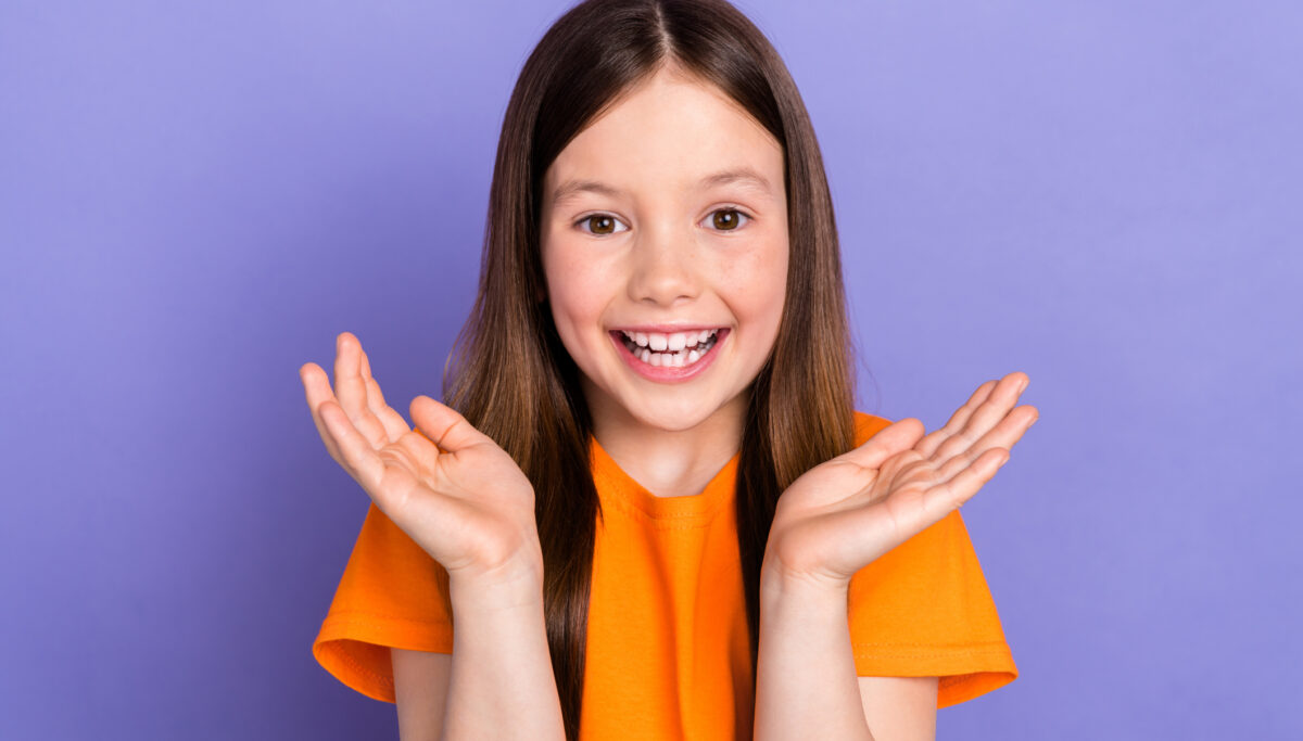 Portrait of young girl preschool age clapping arms positive orange t shirt hooray entertainment day isolated on purple color background.