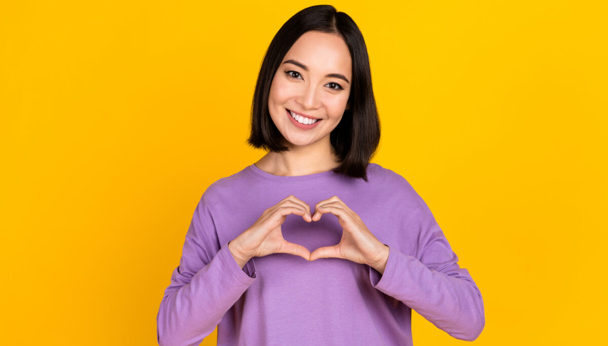 Photo of a happy woman, using her hands to create a heart symbol on a yellow coloured background.
