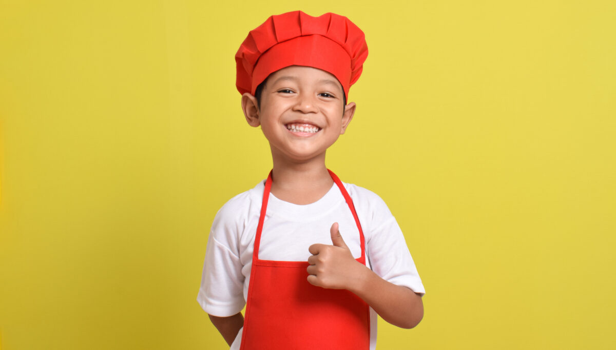 Cute little chef showing thumbs up isolated on yellow background. Asian boy wearing red apron and hat. Great little chef