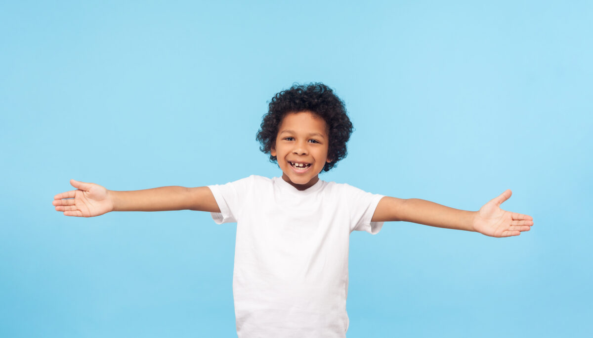 Let's hug. Portrait of friendly hospitable little boy with curls in white T-shirt smiling happily and holding hands wide open to embrace, welcome. indoor studio shot isolated on blue background