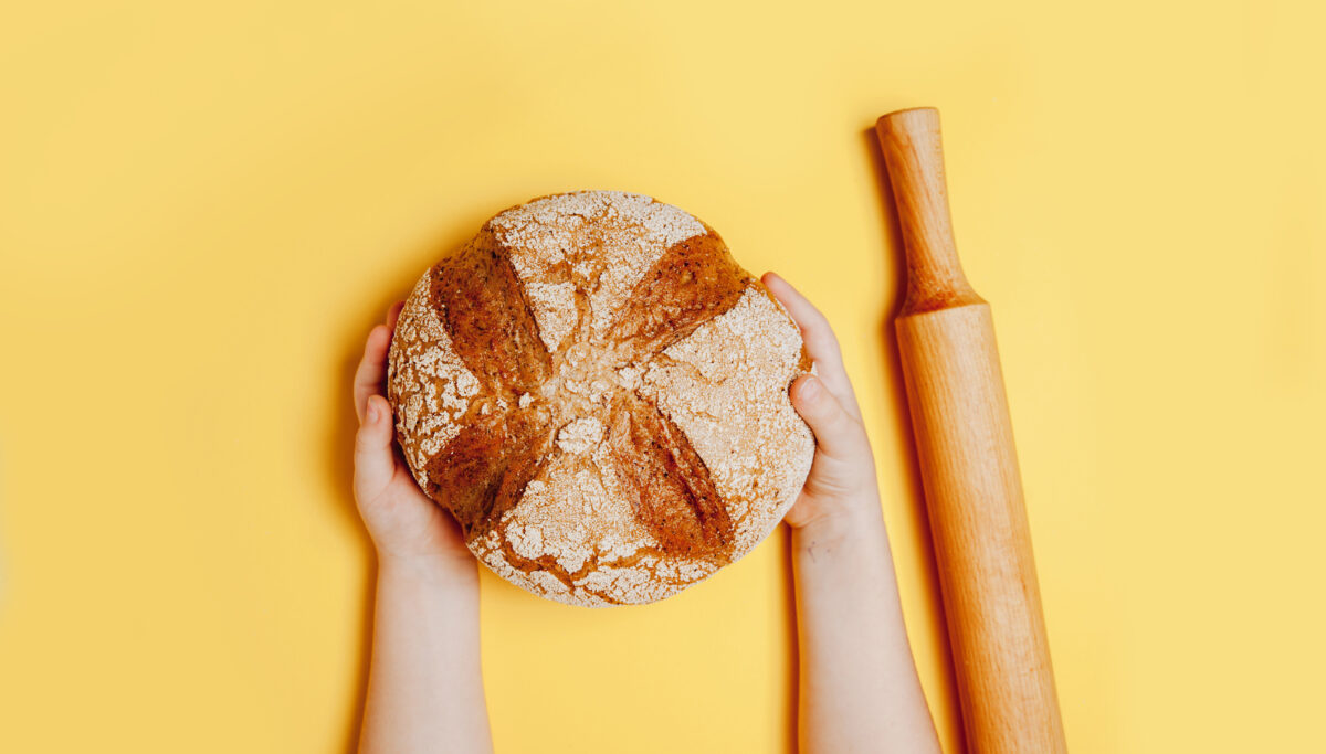 Tasty round bread in child's hands with rolling pin on yellow background.