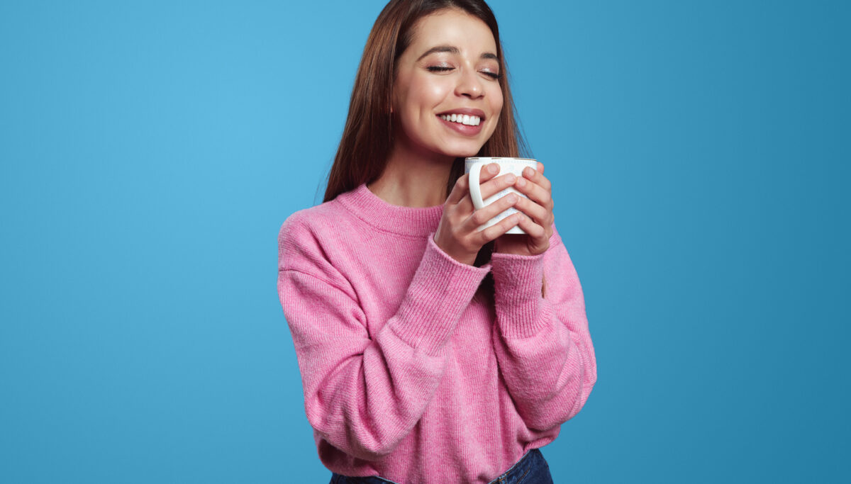 Pretty young woman enjoying drink with closed eyes and holding cup with both hands wearing pink sweater, isolated over blue background