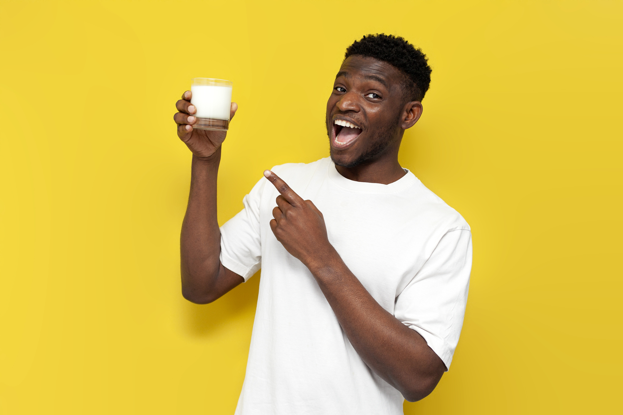 Joyful man holding glass of milk and pointing at it on yellow isolated background.
