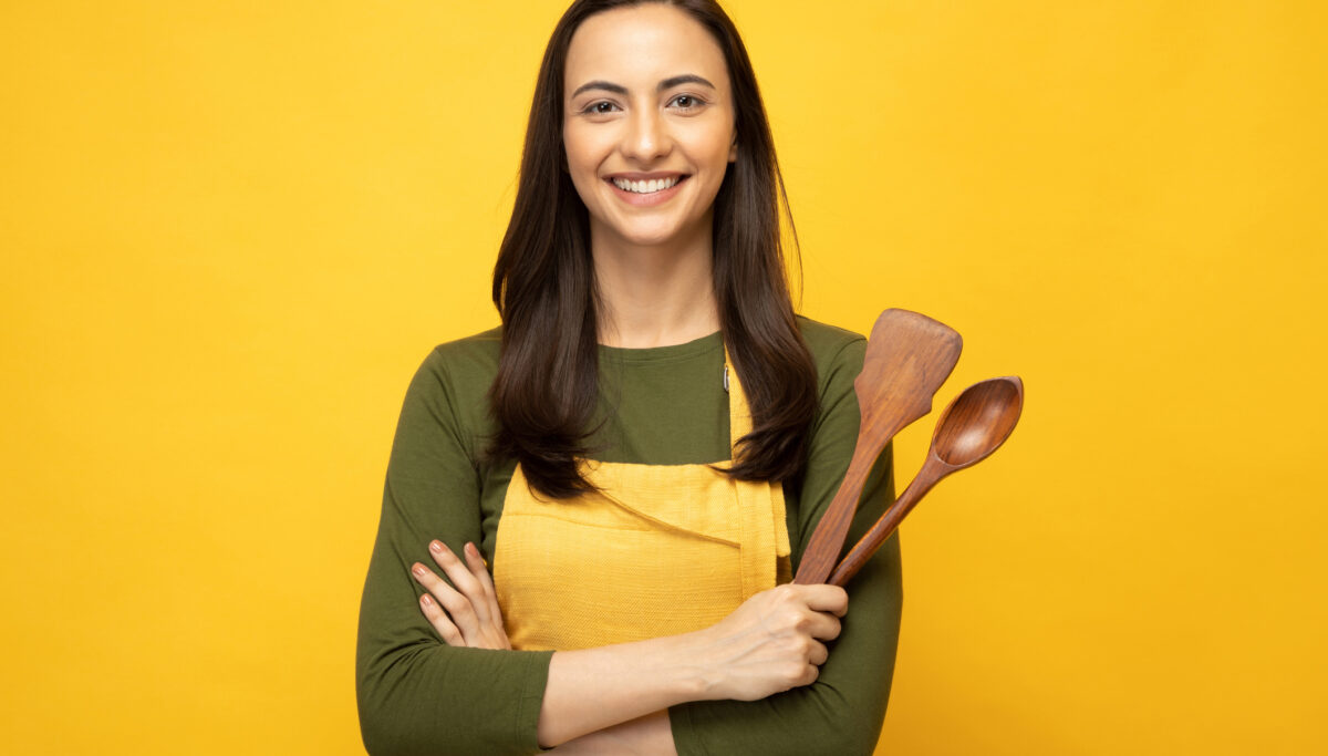 Female chef holding wooden spoon and spatula on a yellow background.