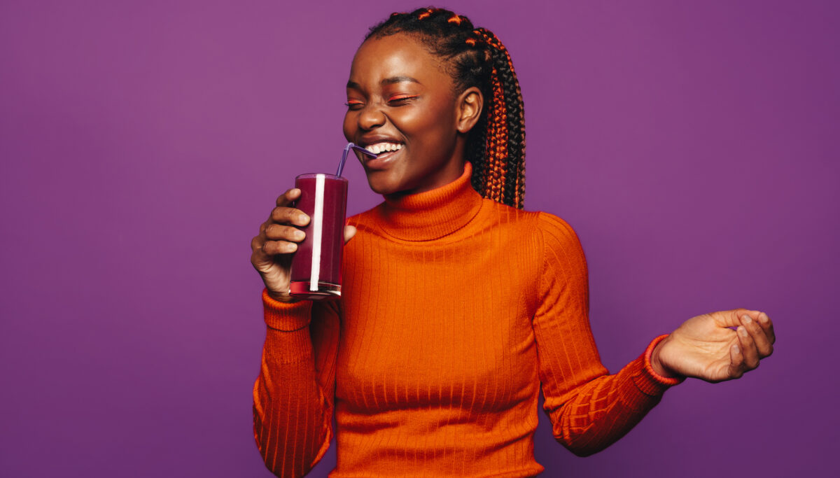 Happy woman with stylish braided hair stands against a purple background, drinking a healthy smoothie.