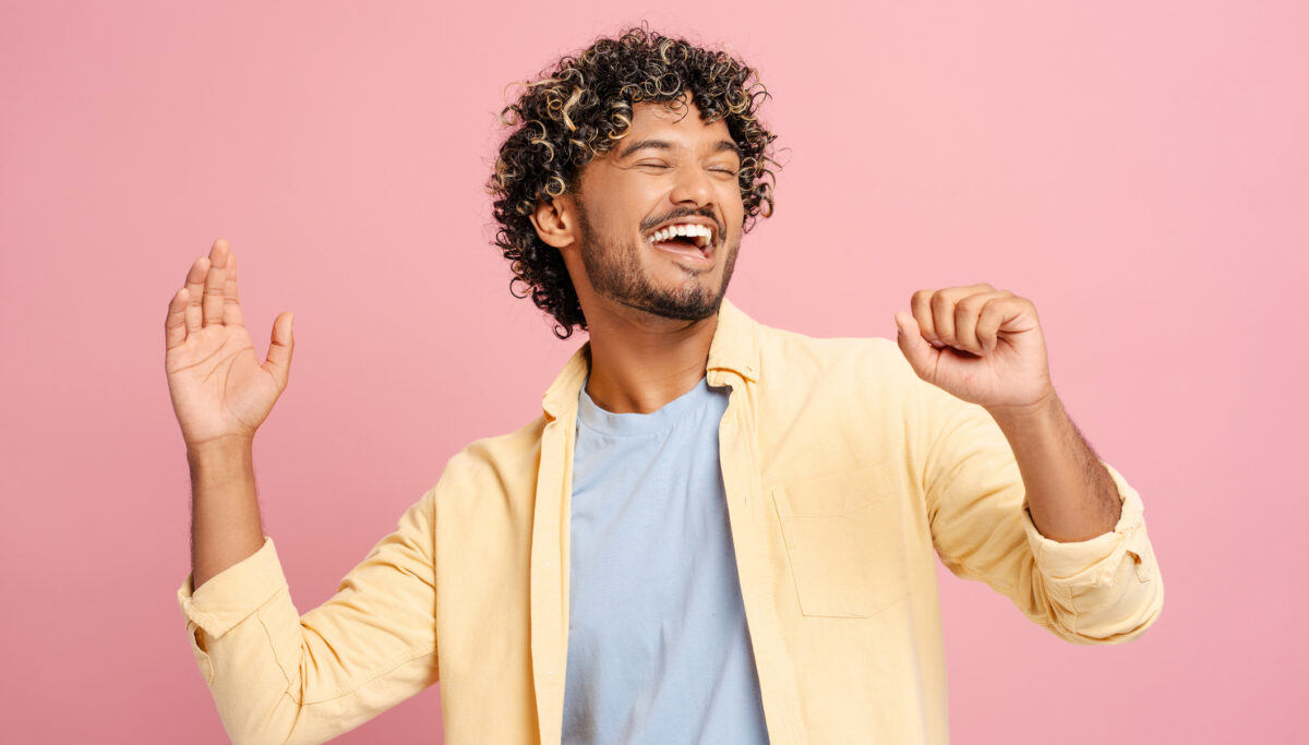 Young man with curly hair dances happily in casual clothes against a pink background, radiating joy and positivity.