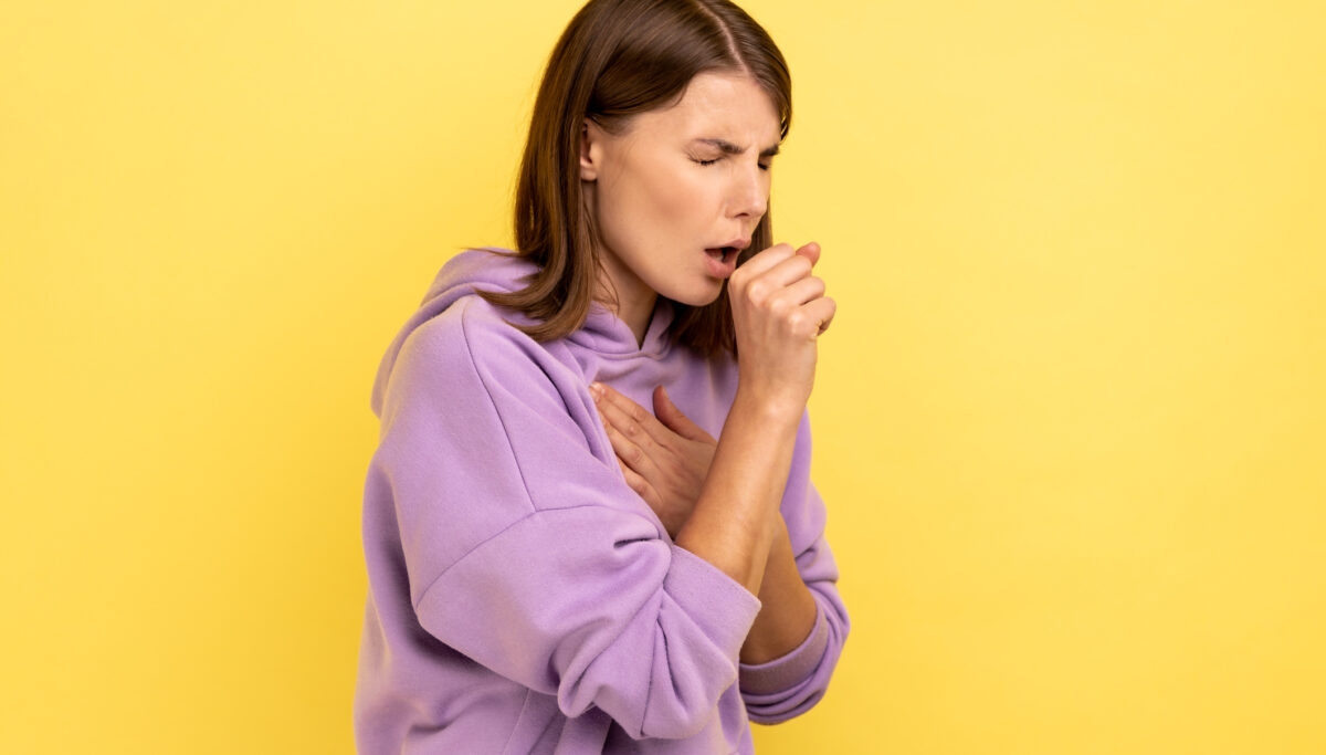 Woman with a cough and hand on her chest, wearing a purple jumper on a yellow background.