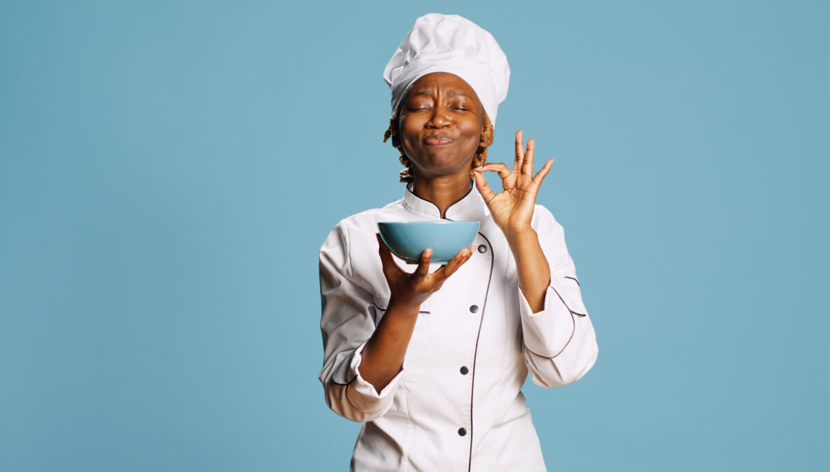 Cheerful gourmet chef smelling food aroma from bowl on camera, enjoying professional meal cooked. Professional female cook savoring tasty food, flavor and scent in studio. Catering service.