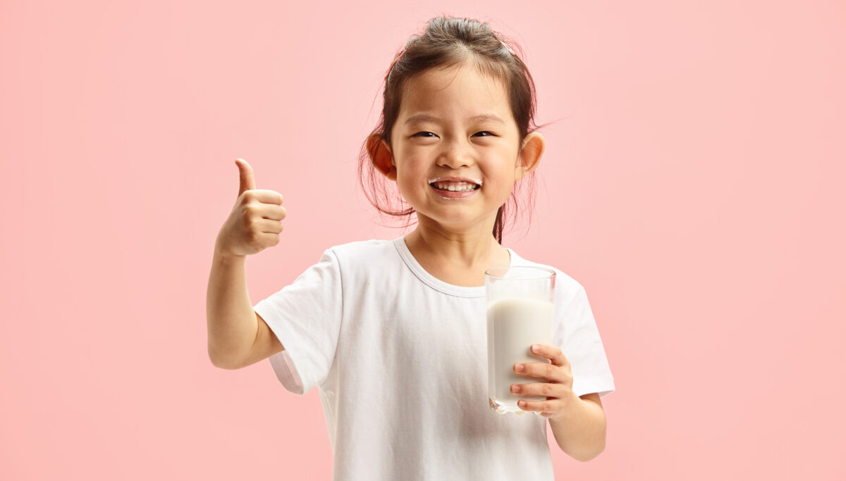 Isolated portrait of cheerful Asian ethnicity little girl holding glass of milk after drinking, rasing thumbup wearinf in white neutral t shirt, expresses positive mood standing over pink isolated background with a free clopy space. Usefulness and natural taste of dairy farm products. Concept of healthy and proper eating.