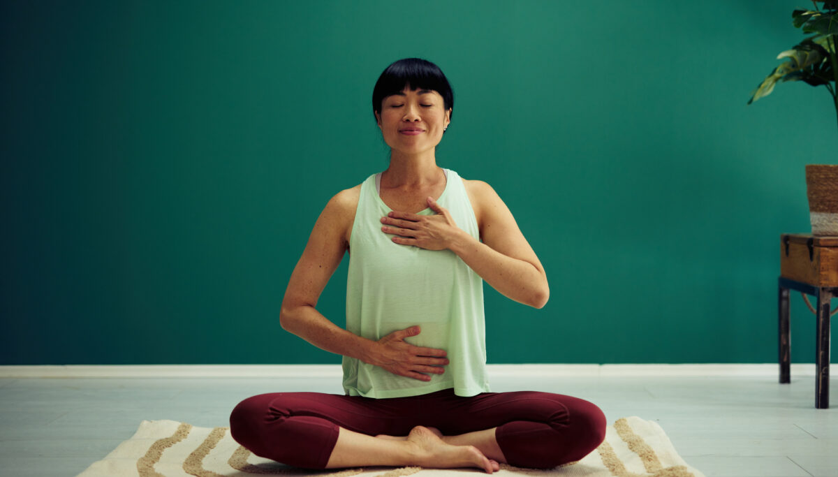 Woman practising yoga, meditating in lotus position with her hands on her chest and belly, focusing on her breathing and mindfulness, finding inner peace and balance