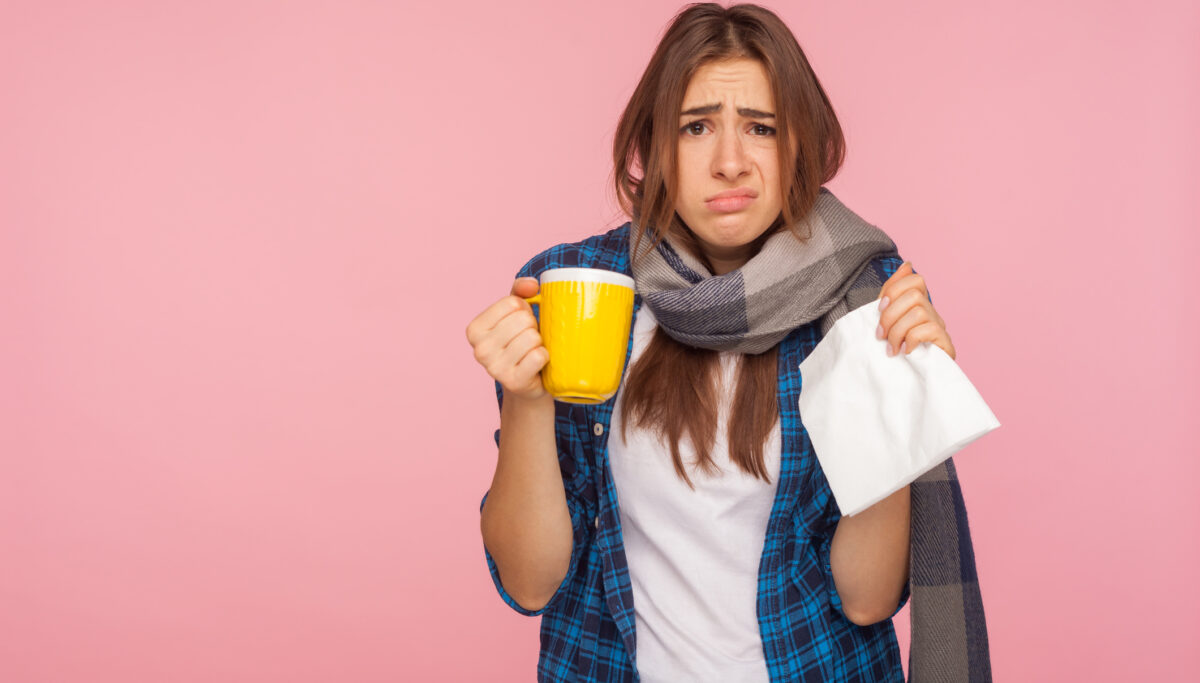 Treatment of seasonal influenza. Portrait of sick girl wrapped in scarf looking with funny displeased grimace, holding tea cup treating flu, cough and fever. studio shot isolated on pink background