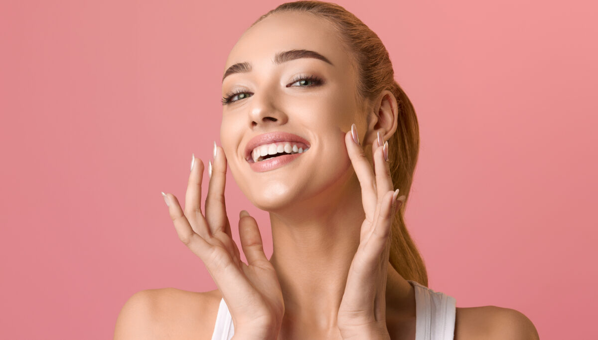 Portrait of beautiful woman touching skin or applying cream over pink background