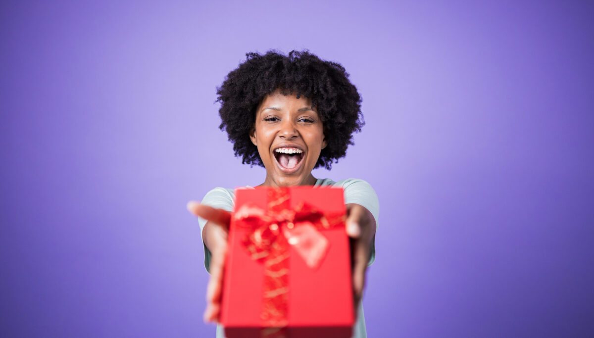Excited young woman giving a red gift box with golden ribbon, expressing joy and generosity on a vibrant purple background