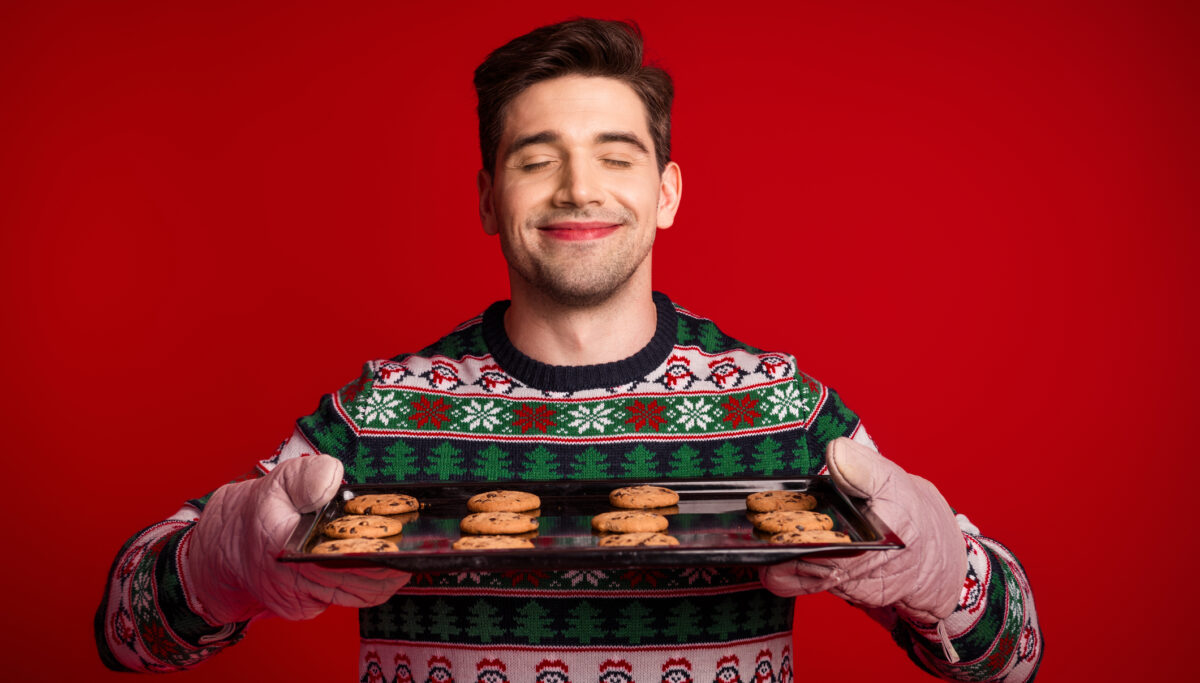 A cheerful man in a festive Christmas sweater presents a tray of cookies against a bold red background. Warm holiday vibes and seasonal celebration with baking and joy.