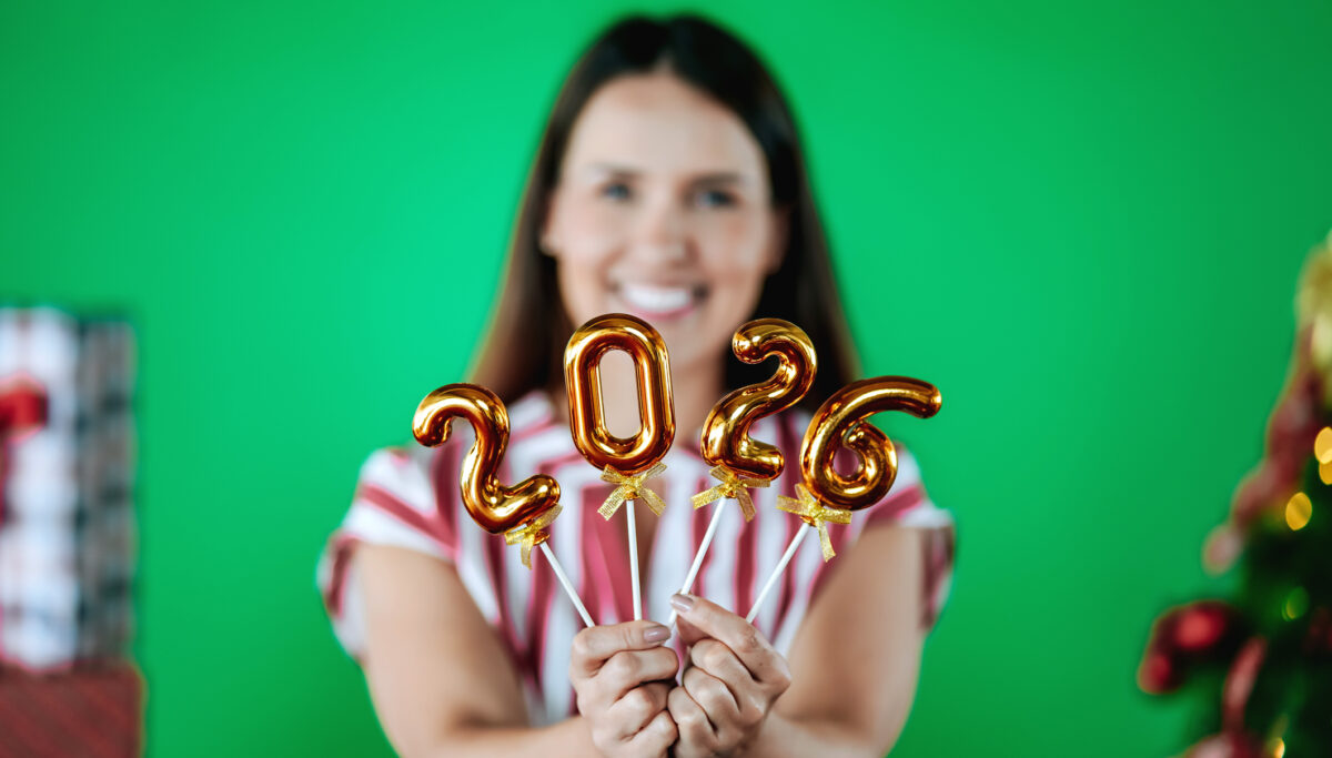 Happy young woman looking directly at the camera and smiling while holding golden balloons forming the number 2026 on a green background.