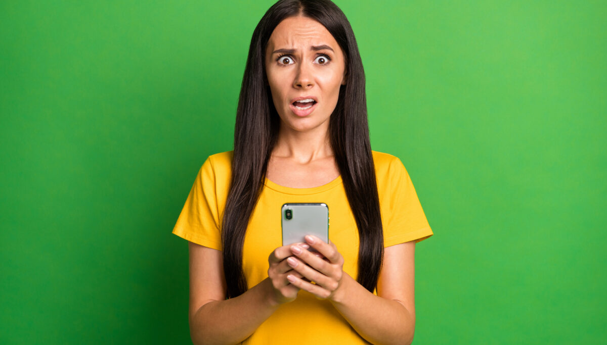 Photo of unhappy scared young woman hold phone dislike reaction addicted blogger isolated on bright green color background