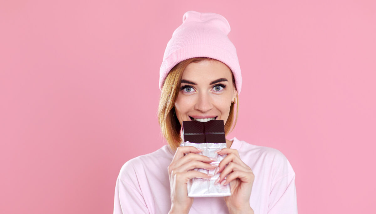 charming happy woman in casual t-shirt biting chocolate bar and looking at camera isolated on pastel pink background