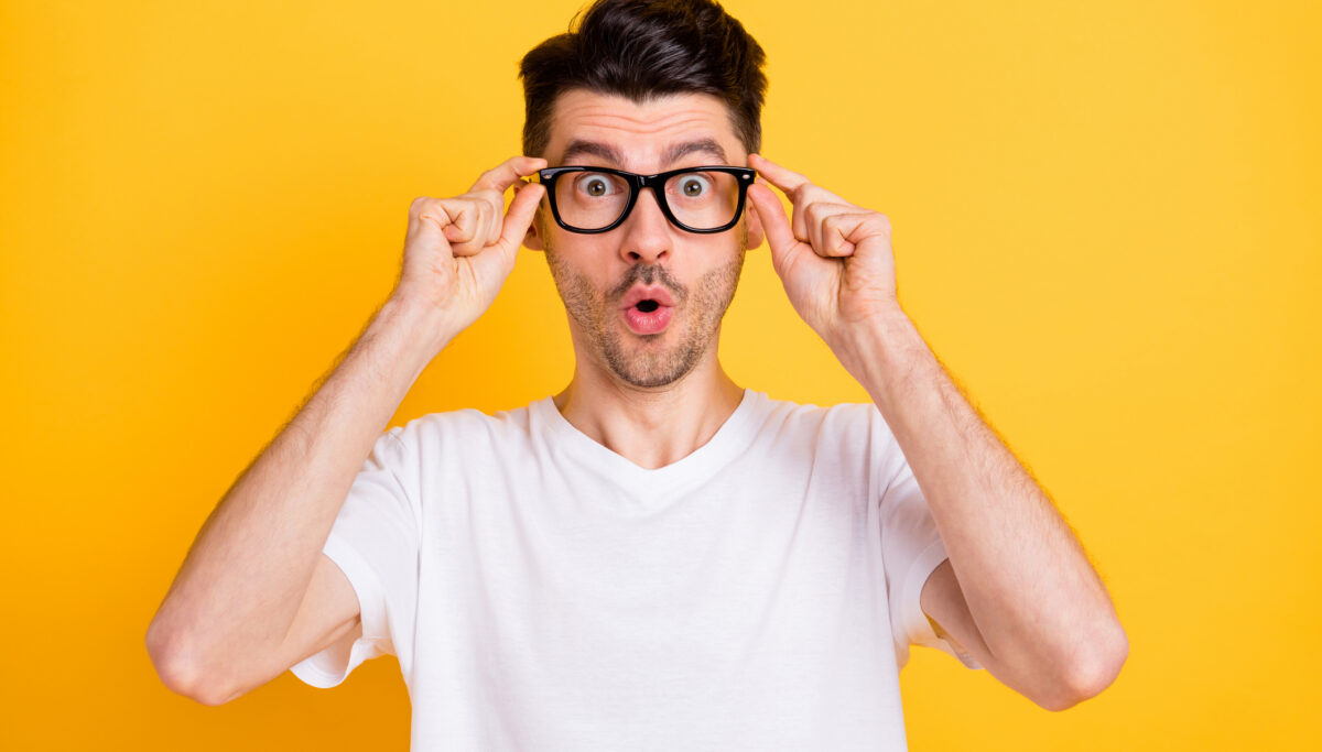 Photo of shocked funny young man wear white t-shirt arms spectacles big eyes isolated yellow color background