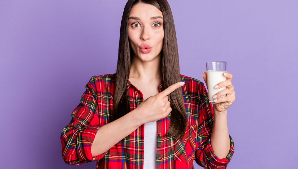 Photo of a woman holding a glass of milk and pointing to it using her other hand, on a purple background.