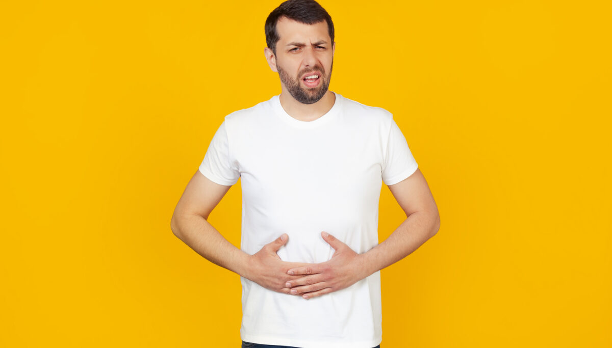 A young man with a beard in a white T-shirt with a hand on his stomach because of indigestion, constipation Stands on isolated yellow background.