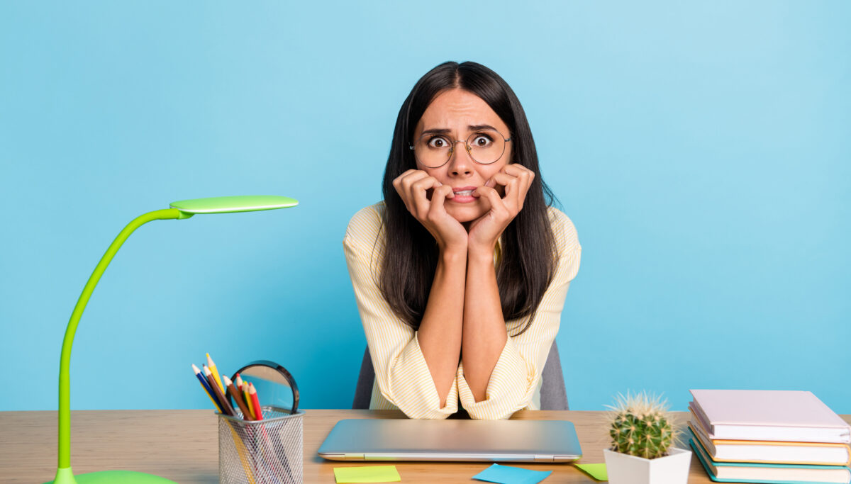 Portrait of stressed lady sitting behind desktop biting finger nail staring camera isolated on blue color background