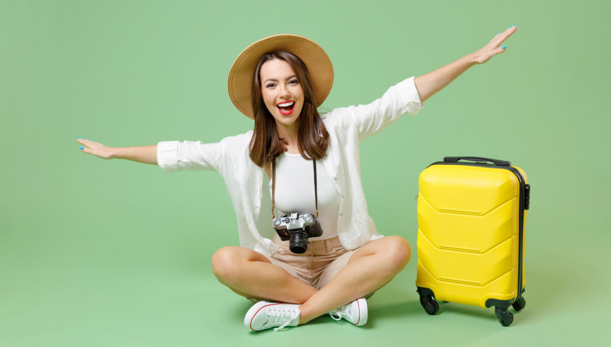 Full length fun happy traveler tourist woman in casual clothes hat suitcase sit with outstretched hands isolated on green background Passenger travel abroad weekend getaway Air flight journey concept